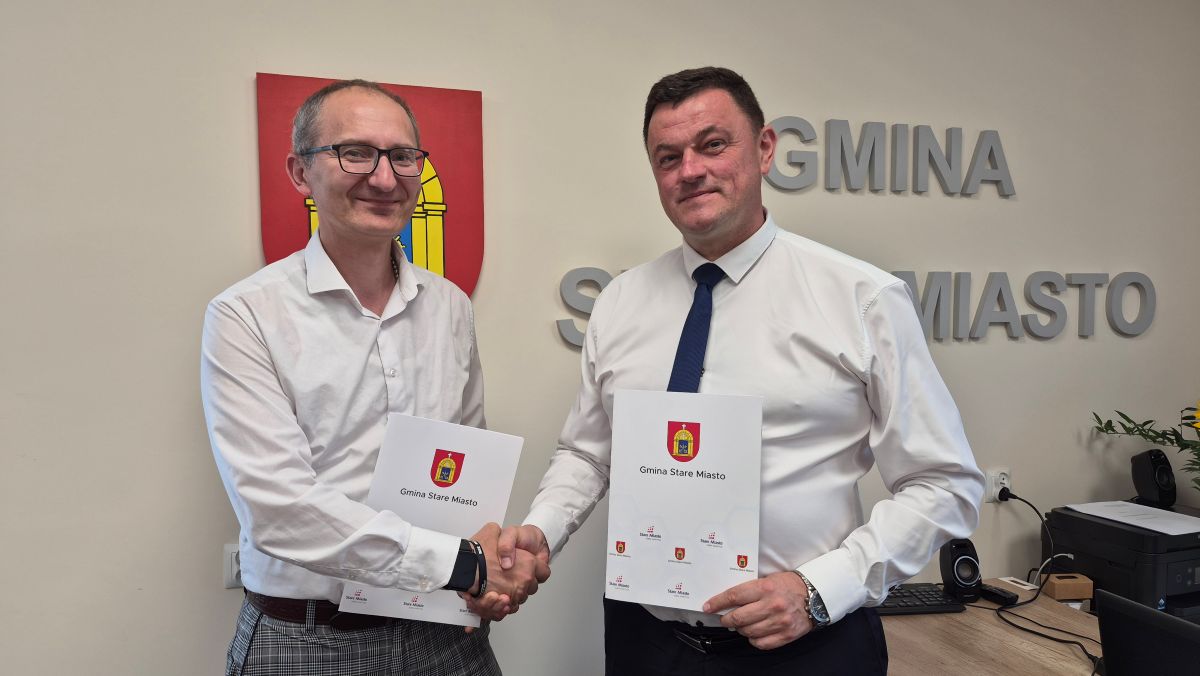 Na zdjęciu dwaj mężczyźni stoją w&nbsp;siedzibie Urzędu Gminy Stare Miasto i&nbsp;podają sobie dłonie po podpisaniu dokument&oacute;w. Każdy z&nbsp;nich trzyma teczkę z&nbsp;herbem i&nbsp;logo Gminy Stare Miasto. W tle widoczne są herb gminy oraz&nbsp;przestrzenny napis &bdquo;Gmina Stare Miasto&rdquo;, a&nbsp;po prawej stronie fragment biurka z&nbsp;wyposażeniem biurowym. Zdjęcie dokumentuje oficjalne spotkanie i&nbsp;zawarcie porozumienia lub&nbsp;podpisanie umowy.