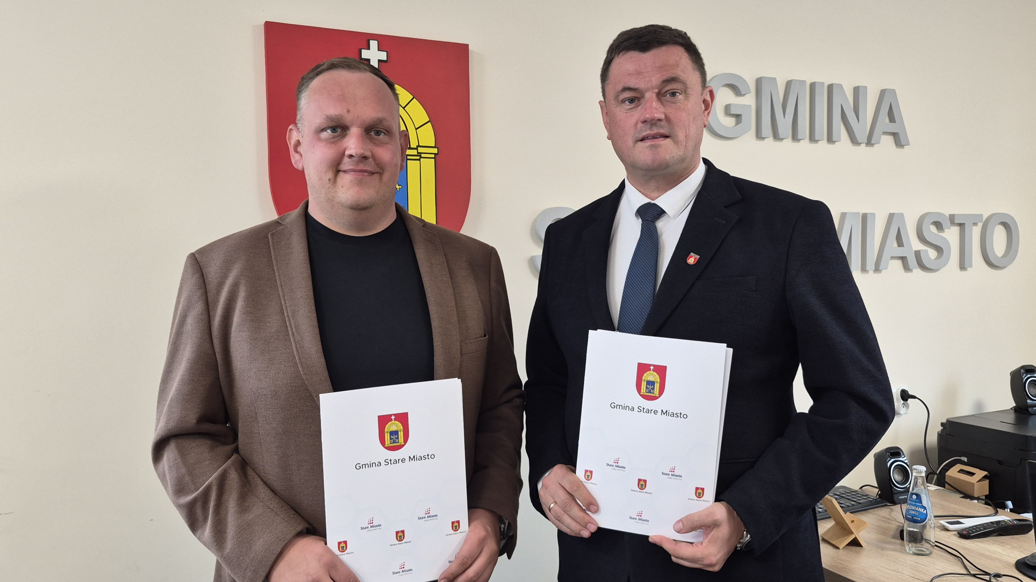 W&oacute;jt Gminy Stare Miasto Dariusz Puchała oraz&nbsp;przedstawiciel firmy KOST-BUD Piotr Druchliński prezentują podpisane dokumenty dotyczące realizacji inwestycji drogowej. Zdjęcie wykonano w&nbsp;Urzędzie Gminy Stare Miasto na&nbsp;tle herbu gminy i&nbsp;napisu &bdquo;Gmina Stare Miasto&rdquo;. Obaj stoją obok siebie, trzymając egzemplarze podpisanej umowy.